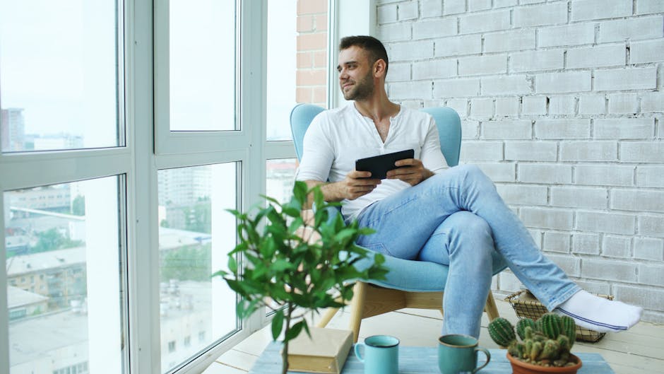 Man seated with tablet near bright window, enjoying leisure time indoors.