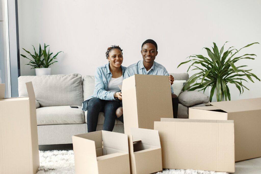 Smiling couple sitting on sofa surrounded by moving boxes in a cozy living room.