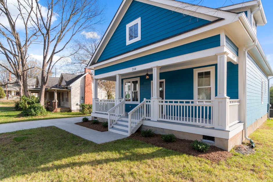 A cozy blue suburban house with a white front porch and lush surroundings on a clear day.